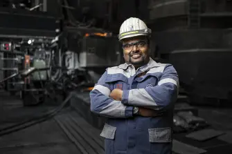 An employee of thyssenkrupp Steel wearing protective clothing and a helmet stands with his arms crossed in an industrial setting. An employee of thyssenkrupp Steel wearing protective clothing and a helmet stands with his arms crossed in an industrial setting.