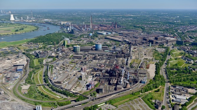 Aerial view of the thyssenkrupp Steel industrial site in Duisburg with production buildings, tanks, smokestacks, and adjacent river infrastructure.