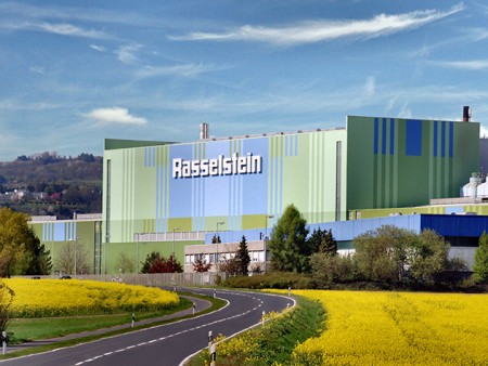 Exterior view of the Packaging Steel plant (thyssenkrupp Rasselstein) with factory buildings, green spaces, and flowering fields. Exterior view of the Packaging Steel plant (thyssenkrupp Rasselstein) with factory buildings, green spaces, and flowering fields.
