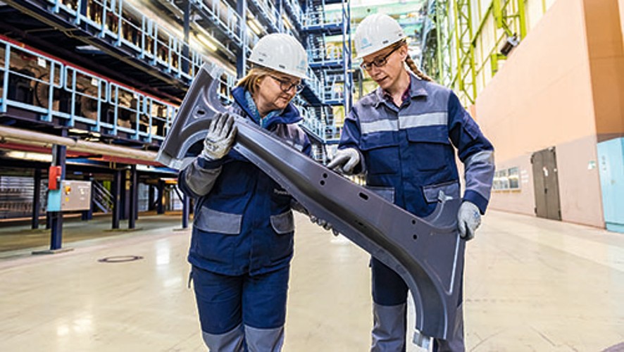 Zwei Mitarbeiterinnen von thyssenkrupp Steel in Schutzkleidung halten gemeinsam ein großes Metallbauteil in einem industriellen Umfeld