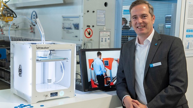 A person in a suit stands next to a 3D printer and a monitor displaying a technical application.