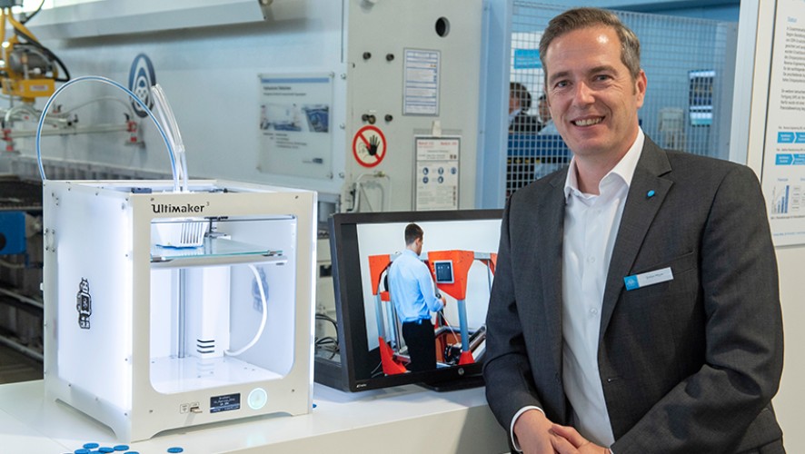A person in a suit stands next to a 3D printer and a monitor displaying a technical application.