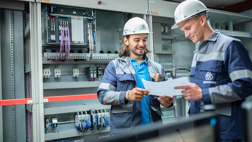 Two thyssenkrupp Steel employees in safety clothing discuss technical details in front of a control panel
