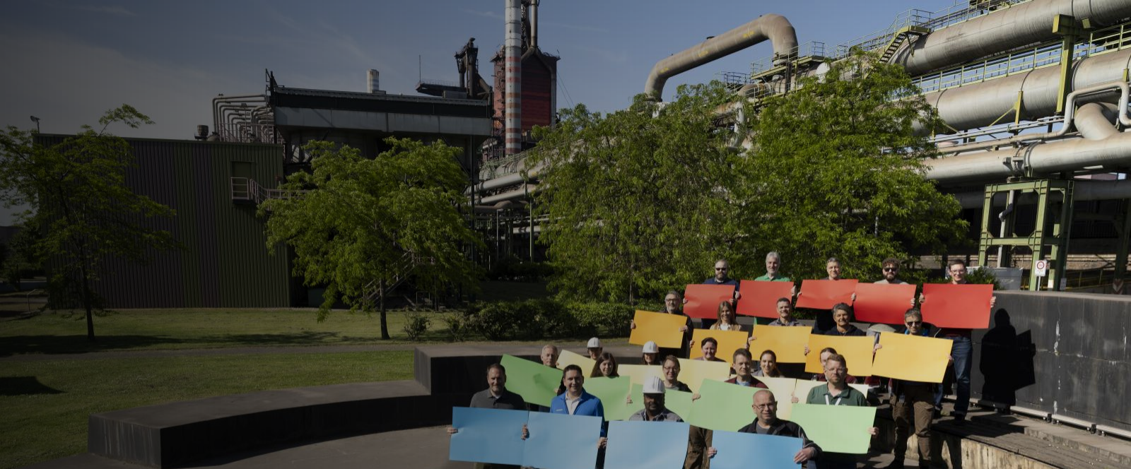 Gruppe von Menschen auf einer Außentreppe bei thyssenkrupp Steel mit farbigen Schildern in Regenbogenanordnung vor industrieller Kulisse und Grünflächen.