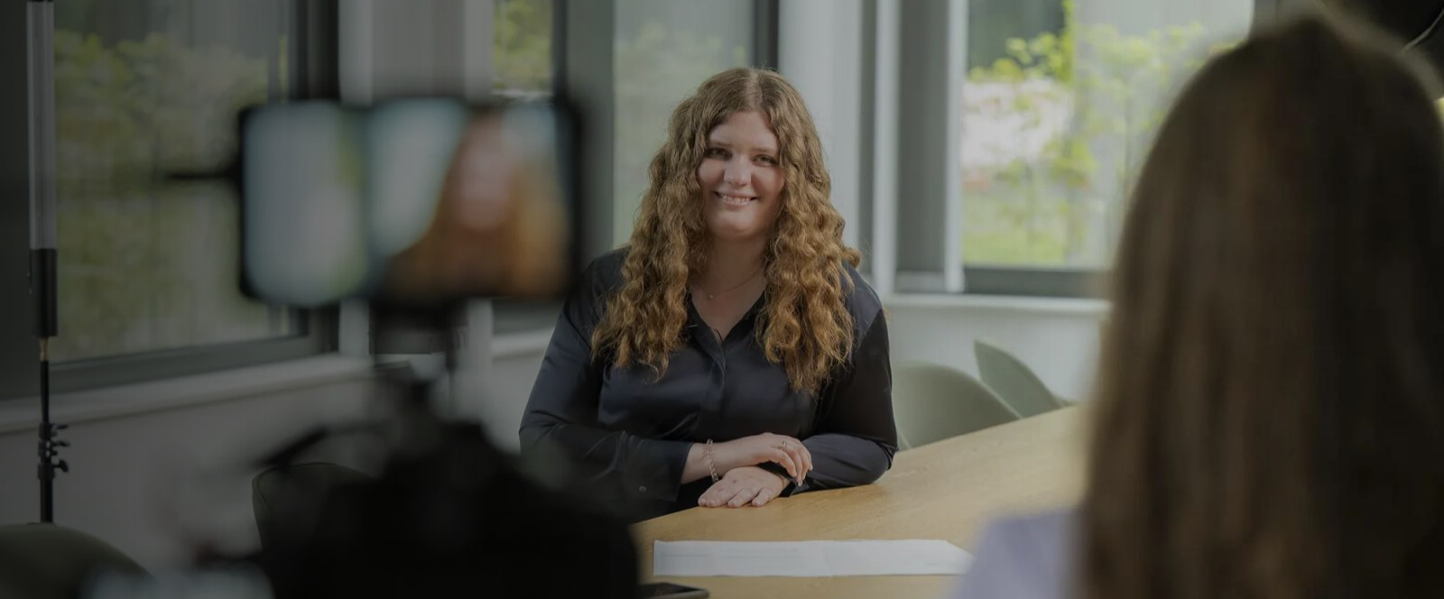 Die interviewte Person ist die Kollegin Pia Fork. Sie sitzt in einem Bürosetting an einem Tisch. Im Vordergrund sind Kameraequipment und eine weitere Person zu sehen. Im Hintergrund große Fenster mit Blick ins Grüne.