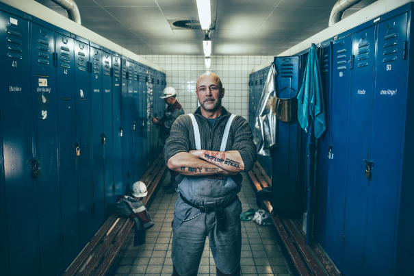 In an industrial locker room, blue lockers are arranged in two rows. In the center stands a person in work clothes with their arms crossed. In the background, you can see more lockers, benches, protective gear, and a helmet.