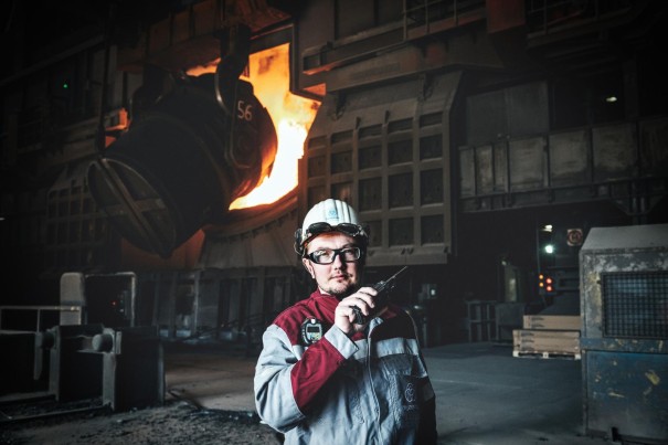 In a large industrial hall, an employee wearing a hard hat and work clothes stands in the foreground. In the background, a production line can be seen, from which molten, glowing steel is flowing.