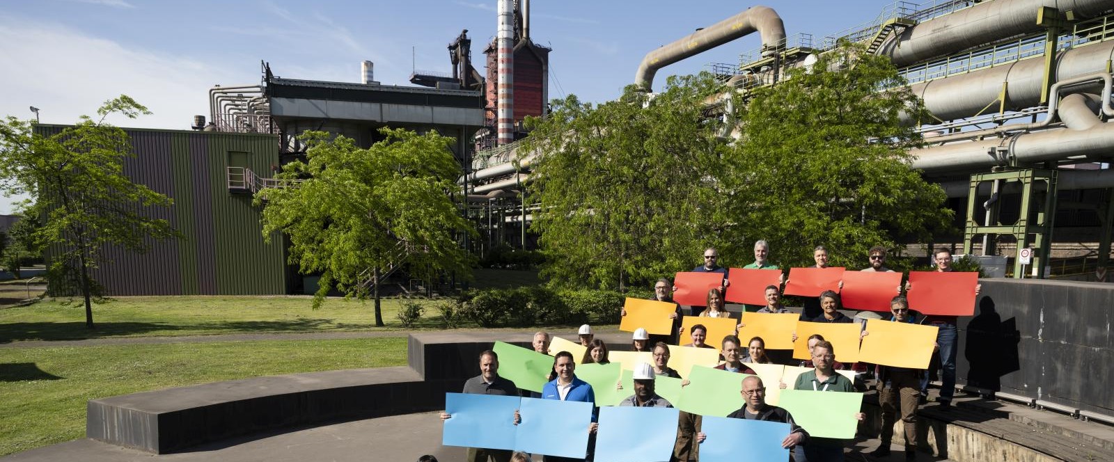 Group of people on an outdoor staircase at thyssenkrupp Steel with colorful signs arranged in a rainbow pattern against an industrial backdrop and green spaces. Group of people on an outdoor staircase at thyssenkrupp Steel with colorful signs arranged in a rainbow pattern against an industrial backdrop and green spaces.
