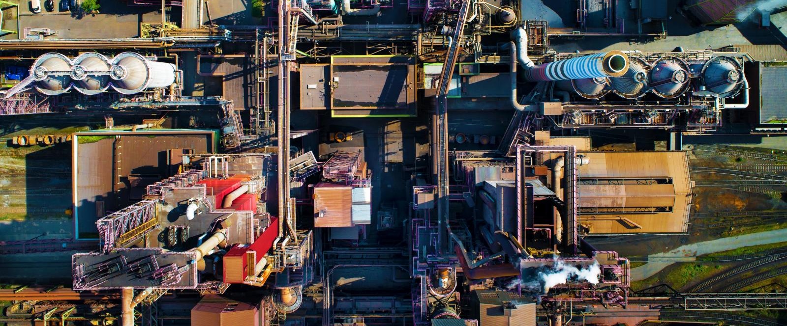 Aerial view of the industrial site in Duisburg with several large buildings, chimneys, storage tanks, and parked vehicles. A road runs along the bottom edge of the image. The facility is divided into different functional areas. Aerial view of the industrial site in Duisburg with several large buildings, chimneys, storage tanks, and parked vehicles. A road runs along the bottom edge of the image. The facility is divided into different functional areas.