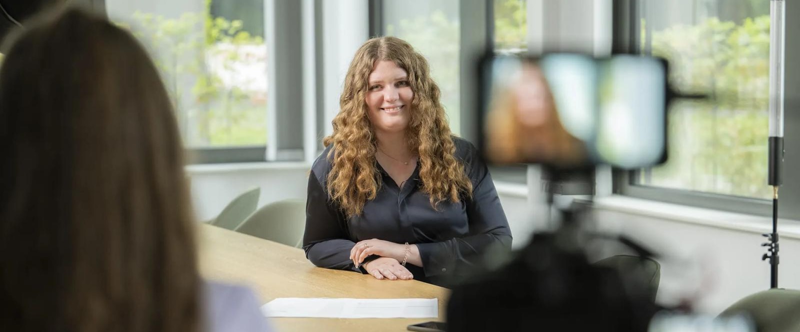 The interviewee is our colleague Pia Fork. She is sitting at a table in an office setting. Camera equipment and another person can be seen in the foreground. In the background are large windows with a view of greenery. The interviewee is our colleague Pia Fork. She is sitting at a table in an office setting. Camera equipment and another person can be seen in the foreground. In the background are large windows with a view of greenery.