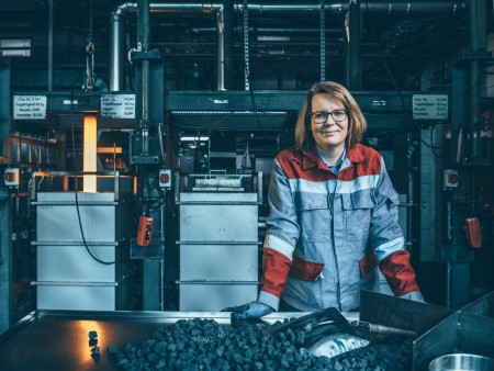 Mitarbeiterin bei thyssenkrupp Steel in rot-grauer Schutzkleidung in einem industriellen Umfeld mit Maschinen und Kohlematerial auf einem Tisch. Mitarbeiterin bei thyssenkrupp Steel in rot-grauer Schutzkleidung in einem industriellen Umfeld mit Maschinen und Kohlematerial auf einem Tisch.