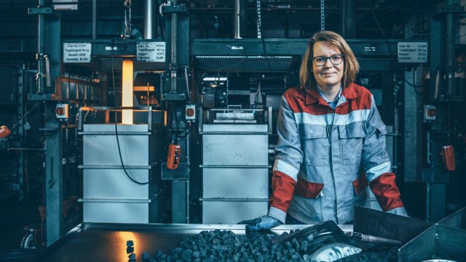 An employee at thyssenkrupp Steel wearing red-and-gray protective clothing in an industrial setting with machinery and coal on a table. 