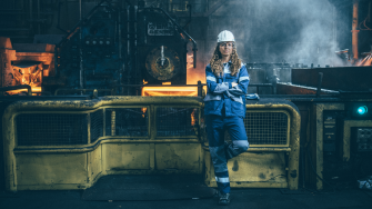 Employee at thyssenkrupp Steel wearing blue workwear and a white helmet, standing in front of yellow safety barriers in an active industrial environment with machinery.