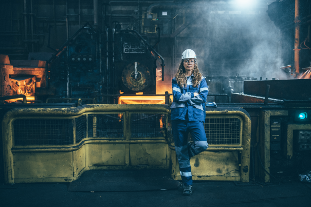 In a large industrial hall, a female employee wearing blue work clothes and a hard hat stands in front of a machine processing red-hot steel. Railings, machines, and pipes surround the work area. 