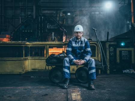 A thyssenkrupp Steel employee wearing blue and white striped work clothes and a white helmet in a factory with machinery and safety barriers. A thyssenkrupp Steel employee wearing blue and white striped work clothes and a white helmet in a factory with machinery and safety barriers.