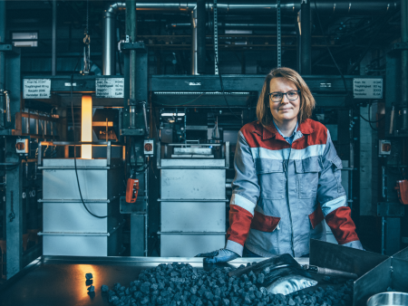 Employee at thyssenkrupp Steel wearing red and gray protective clothing in an industrial environment with machines and coal material on a table. Employee at thyssenkrupp Steel wearing red and gray protective clothing in an industrial environment with machines and coal material on a table.