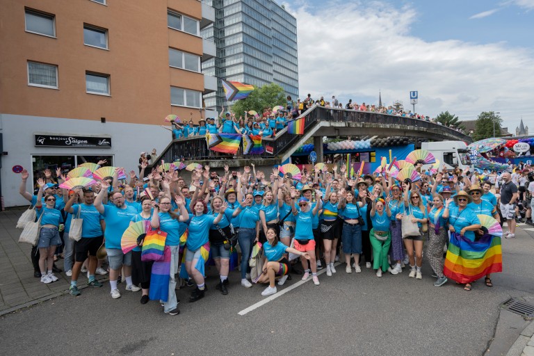 thyssenkrupp employees take part in Cologne Pride (CSD), wearing blue shirts and carrying rainbow flags and fans. Spectators are visible on an elevated platform in the background. thyssenkrupp employees take part in Cologne Pride (CSD), wearing blue shirts and carrying rainbow flags and fans. Spectators are visible on an elevated platform in the background.