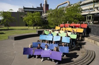 Eine Gruppe von thyssenkrupp Steel Mitarbeitenden sitzt auf Außentreppen und hält große Farbtafeln in einem Farbverlauf von Lila bis Rot. Im Hintergrund sind Bäume und industrielle Strukturen zu sehen.