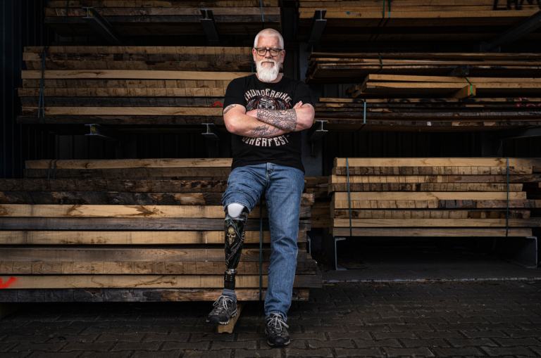 A man with a prosthetic leg leans against stacked wooden planks in a lumberyard. He wears a black T-shirt, jeans, and black shoes. A man with a prosthetic leg leans against stacked wooden planks in a lumberyard. He wears a black T-shirt, jeans, and black shoes.